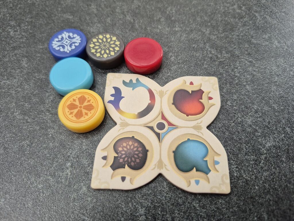 Neben einem Plättchen liegen runde Spielsteine in fünf verschiedenen Farben.