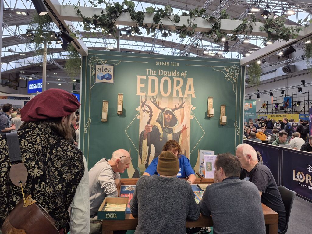 Eine Werbewand von "The Druids of Edora", davor sitzen Menschen an einem Spieltisch.