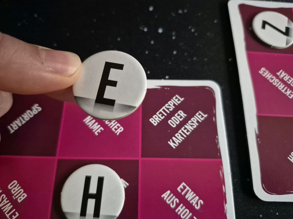 Eine Hand hält ein Plättchen mit einem E über einen Spielplan.