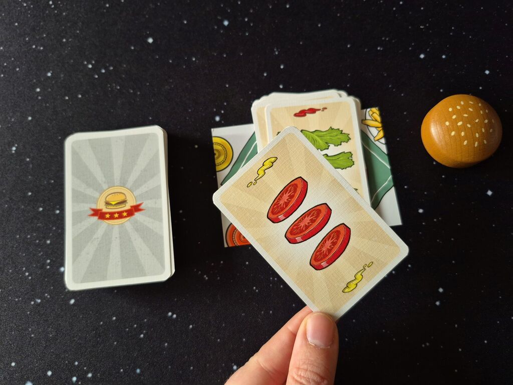 Eine Hand hälte eine Karte über das restliche Spielmaterial von "Burger Slam".