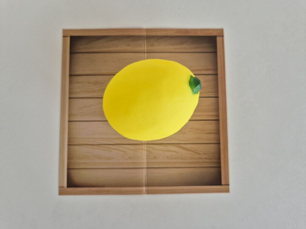 Eine knallgelbe Spielzeugzitrone in einer Schachtel mit Holzoptik "Easy Peasy Lemon Squeaky".