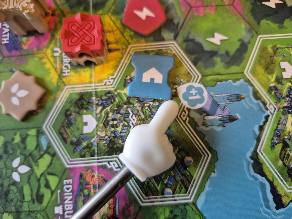 Ein Zeiger in Form eines Comicfingers zeigt auf ein Plättchen auf dem Spielplan.