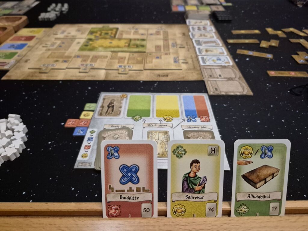 Drei Karten in einer Halterung, dahinter Spielmaterial.