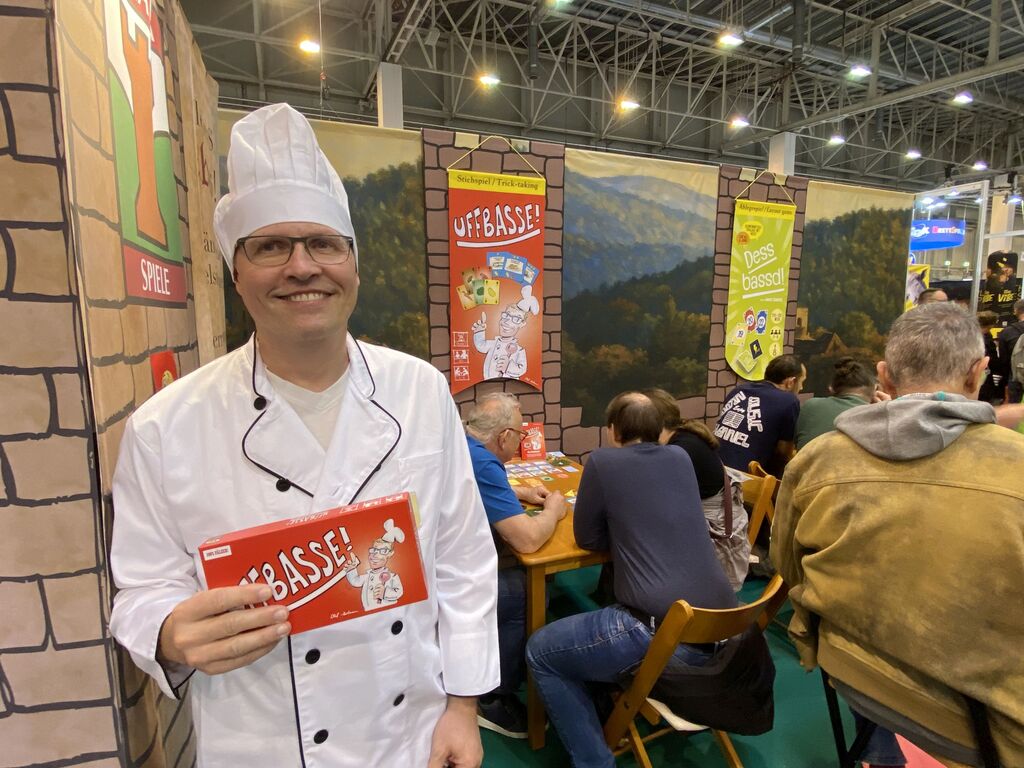 Ein Mann in Kochjacke hält ein Spiel an einem Messestand.