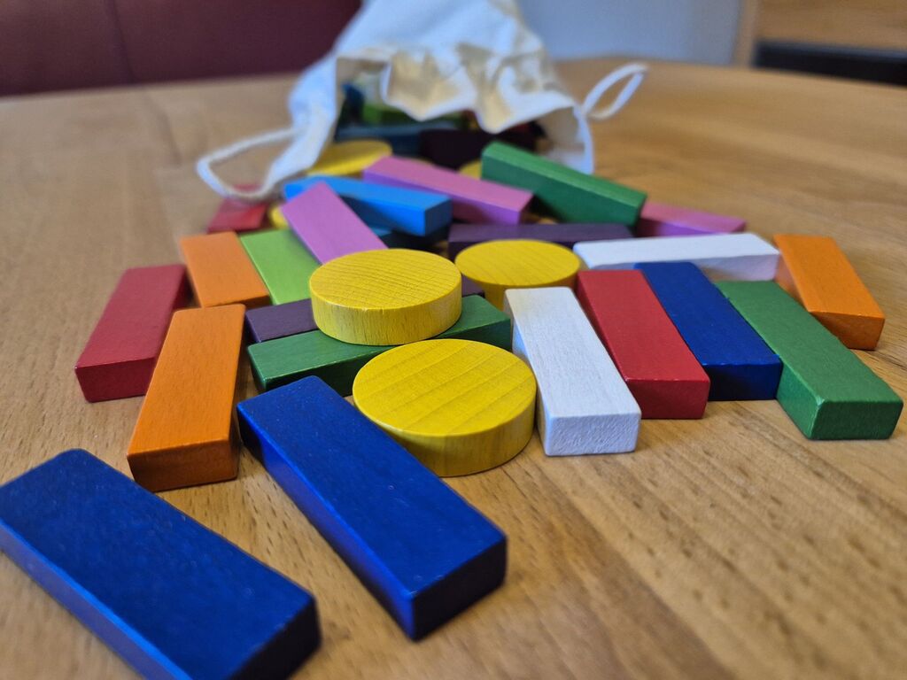 Rechteckige bunte Holzspielsteine, gelbe Holzscheiben, dahinter ein heller Beutel.